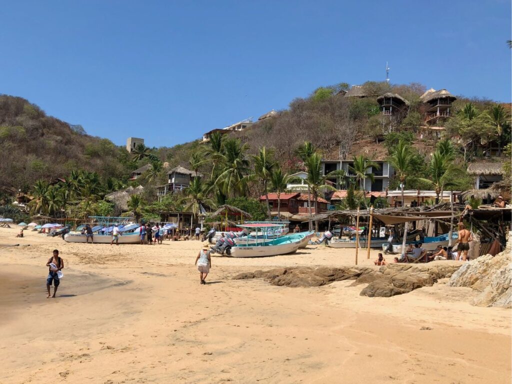 beach shacks, boats, and travellers walking on beach in mazunte, oaxaca